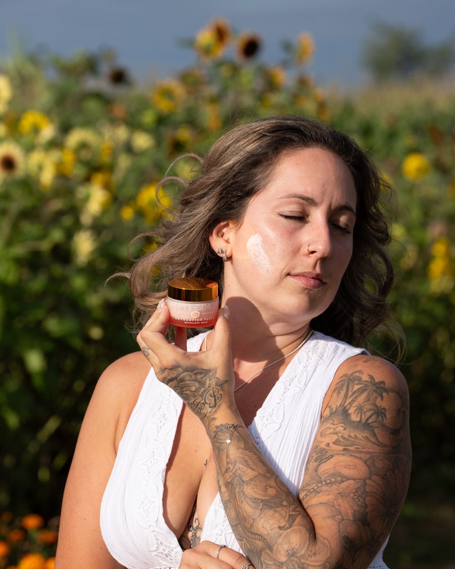 Une femme applique une crème B&F Essence sur sa joue, les yeux fermés, dans un jardin
