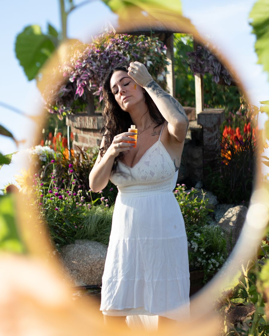 Reflet, dans un miroir rond tenu parmi les tournesols, d'une femme en robe blanche qui tient la lotion tonique Lotus Solaire